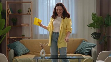 Günlük yaşam tarzı konsepti. Genç bir kadın evde vakit geçiriyor, hafta sonları temizlikçi kadın oturma odasında dans ediyor ve masadaki tozları siliyor..
