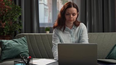 Günlük yaşam tarzı konsepti. Genç, yetişkin bir iş kadını, oturma odasında oturmuş dizüstü bilgisayarla çalışıyor, not defterini kontrol ediyor ve ekrana bakıyor..
