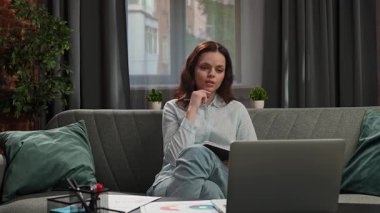Günlük yaşam tarzı konsepti. Genç, yetişkin iş kadını ev ofisinde vakit geçiriyor. Oturma odasında oturan, dizüstü bilgisayarda çalışan, internetten video izleyen ve not defterine yazan bir kadın..