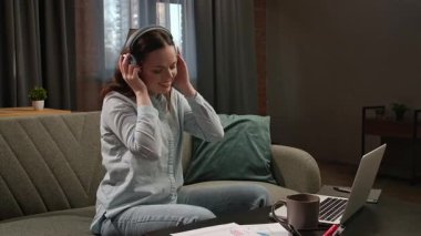 Günlük yaşam tarzı konsepti. Genç, yetişkin bir iş kadını, oturma odasında oturmuş laptopla çalışıyor, kulaklıkla müzik dinliyor ve dans ediyor..