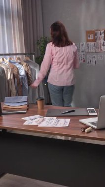 Günlük yaşam tarzı konsepti. Genç yetişkin iş kadını ofiste vakit geçiriyor, kadın moda tasarımcısı atölyede çalışıyor, kahve içiyor, şık kıyafetlerle askılara bakıyor..