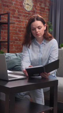 Günlük yaşam tarzı konsepti. Genç yetişkin iş kadını ev ofisinde vakit geçiriyor. Oturma odasında oturan kadın dizüstü bilgisayarda çalışıyor. Hesaplamalar yapıyor ve deftere yazıyor..