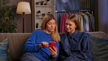 İki kadın bir kanepede oturuyor, akıllı bir telefonla meşgul, rahat bir oturma odasında keyifli anlar paylaşıyorlar..