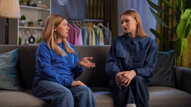 İki kadın bir kanepede oturuyor, derin bir tartışma içindeler. Arka planda çeşitli günlük giysi öğeleri yer alıyor.