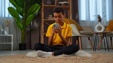 Günlük yaşam tarzı konsepti. Genç Afrikalı Amerikalı çocuk evde boş zaman geçiriyor, çocuk oturma odasında yerde oturuyor ve akıllı telefondan internete mesaj atıyor..