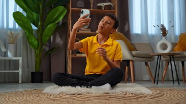 Günlük yaşam tarzı konsepti. Genç Afrikalı Amerikalı çocuk evde boş zaman geçiriyor, çocuk oturma odasında yerde oturuyor ve akıllı telefondan komik selfie çekiyor..
