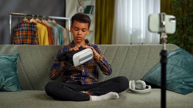 Günlük yaşam tarzı konsepti. Genç Afrikalı Amerikalı çocuk evde vakit geçiriyor, çocuk blogcu koltukta oturuyor akıllı telefondaki videoları kaydediyor, kameraya konuşuyor ve video gözlüklerini gösteriyor ve eleştiri yapıyor..