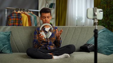 Günlük yaşam tarzı konsepti. Genç Afrikalı Amerikalı çocuk evde vakit geçiriyor, çocuk blogcu koltukta oturuyor akıllı telefondaki videoları kaydediyor, kameraya konuşuyor kulaklıkları gösteriyor ve eleştiri yapıyor..
