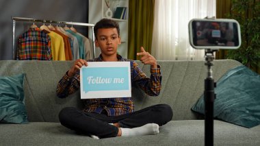 Günlük yaşam tarzı konsepti. Genç Afrikalı Amerikalı çocuk evde vakit geçiriyor, çocuk blogcu koltukta oturuyor akıllı telefondaki videoları kaydediyor, kamera karşısında konuşuyor kağıt sayfalarda benimle mesajlaşıyor..