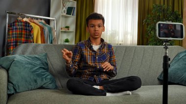 Günlük yaşam tarzı konsepti. Genç Afrikalı Amerikalı çocuk evde vakit geçiriyor, çocuk blogcu koltukta oturuyor akıllı telefondaki videoları kaydediyor, kameraya konuşuyor, hikayeler ve şakalar paylaşıyor..