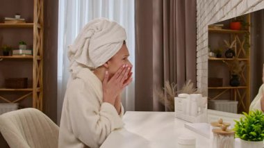 Kafasında havlu olan bornoz giymiş bir kadın aydınlık bir odada ayna ile cildini yakından inceliyor..