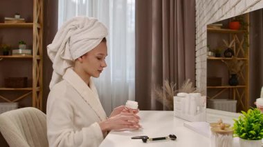 Kafasında havlu olan bir kadın makyaj aynasına krem sürüyor, rutin cilt bakımına odaklanıyor..