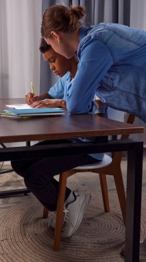 Günlük yaşam tarzı konsepti. Genç Afrikalı Amerikalı çocuk evde okuyor, çocuk ve anne masada, çocuk ödevlerini deftere yazıyor, anne yardım ediyor ve kontrol ediyor..