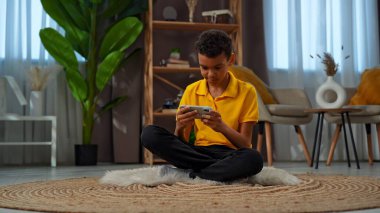 Günlük yaşam tarzı konsepti. Genç Afrikalı Amerikalı çocuk evde boş zaman geçiriyor, çocuk oturma odasında yerde oturuyor ve akıllı telefondan video oyunu oynuyor..