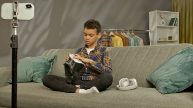 Günlük yaşam tarzı konsepti. Genç Afrikalı Amerikalı çocuk evde vakit geçiriyor, çocuk blogcu koltukta oturuyor akıllı telefondaki videoları kaydediyor, kameraya konuşuyor ve video gözlüklerini gösteriyor ve eleştiri yapıyor..