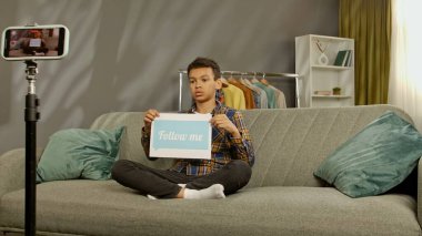 Günlük yaşam tarzı konsepti. Genç Afrikalı Amerikalı çocuk evde vakit geçiriyor, çocuk blogcu koltukta oturuyor akıllı telefondaki videoları kaydediyor, kamera karşısında konuşuyor kağıt sayfalarda benimle mesajlaşıyor..