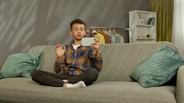 Günlük yaşam tarzı konsepti. Genç Afrikalı Amerikalı çocuk evde boş zaman geçiriyor, çocuk kanepede oturuyor ve akıllı telefondan internetten konuşuyor..