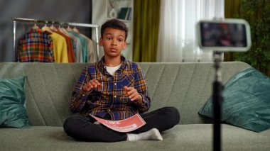 Günlük yaşam tarzı konsepti. Genç Afro-Amerikan çocuk evde vakit geçiriyor, çocuk blogcu akıllı telefondan video kaydediyor, kamera önünde konuşuyor kağıt kağıtları gösteriyor ve abone oluyor. Yavaş çekim.