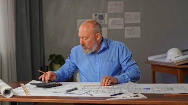 Mavi gömlekli bir mimar hesap makinesi kullanarak hesaplama yapar ve bilgiyi bir deftere yazar. Evin ve dairenin planları masaya serildi..