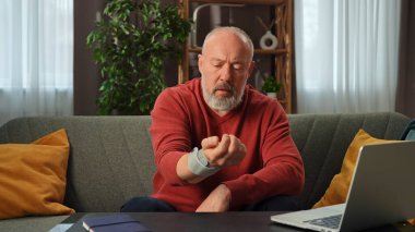 Kırmızı kazaklı yaşlı bir adam dizüstü bilgisayarla çevrili bir kanepede otururken kan basıncını bir tonometreyle ölçer..