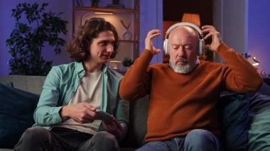 Genç adam akıllı telefon kullanarak kulaklıkla müzik dinlemeyi gösteriyor. Yaşlı erkek büyükbaba akşam oturma odasında torunuyla kanepede oturuyor. Farklı nesil ve öğrenim.