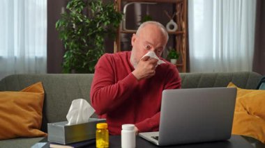 Kırmızı kazaklı yaşlı bir adam kanepede otururken burnunu siliyor. Etrafı ilaç şişeleri, dizüstü bilgisayar ve peçete kutusuyla çevrili..