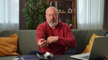 Kırmızı kazaklı yaşlı bir adam dizüstü bilgisayarla çevrili bir kanepede otururken kan basıncını bir tonometreyle ölçer..
