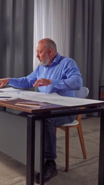 Mavi gömlekli bir mimar planlardaki mesafeleri ölçmek için daireler kullanıyor. Evlerin ve dairelerin yerleşim planlarını masanın üzerine yaydık. Dikey video