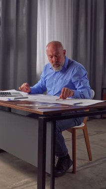 Mavi gömlekli bir mimar dizüstü bilgisayarda klavye kullanıyor. Evlerin ve dairelerin yerleşim planlarının olduğu çizimler masanın üzerine yayılmış. Dikey video