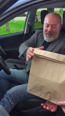 Yaşlı bir adam arabada oturuyor ve paket servis poşetine bakıyor. Yemek dağıtımını gösteriyor. Dikey video