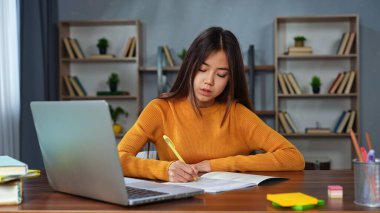 Sarı kazaklı genç bir kız masasında dizüstü bilgisayar kullanırken not defterine notlar yazmaya konsantre oluyor..