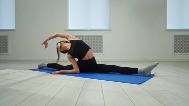 Bir kadın, geniş, aydınlık bir odada mavi bir paspasın üzerinde bacaklarını ayırarak esneklik ve dengeyi gösterir..