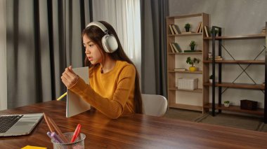 Kitap ve bitkilerle dolu rahat bir ev ortamında kulaklık takarak derslere odaklanan genç bir kız..