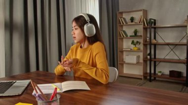 Kulaklık takan genç bir kadın, yakınlarda bir dizüstü bilgisayar ve açık defterlerle ahşap bir masada ders çalışıyor..