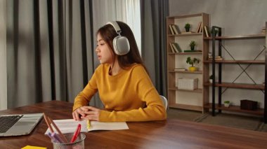 Kulaklık takan genç bir kadın, yakınlarda bir dizüstü bilgisayar ve açık defterlerle ahşap bir masada ders çalışıyor..