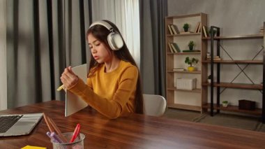 Kitap ve bitkilerle dolu rahat bir ev ortamında kulaklık takarak derslere odaklanan genç bir kız..