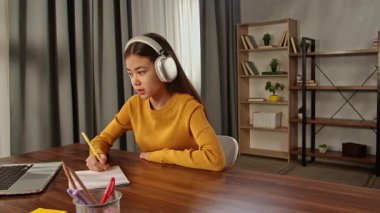 Kulaklıklı sarı kazaklı genç bir kız, dizüstü bilgisayarını online ders için kullanırken not defterine dikkatlice notlar yazar..