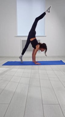 Genç bir kadın, mavi bir mindere karmaşık bir yoga pozu veriyor, dayanıklılık ve esneklik sergiliyor. Dikey video