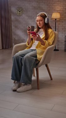 Günlük yaşam tarzı konsepti. Genç kız akşam vaktini evde geçiriyor, kız oturma odasında oturmuş çay içiyor ve akıllı telefonuyla mesajlaşıyor, kulaklık takıyor. Dikey video