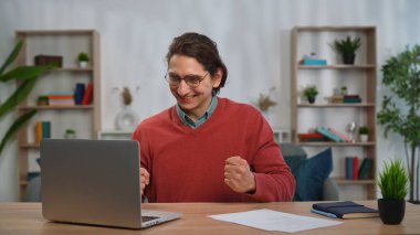 Günlük yaşam tarzı konsepti. Genç yetişkin bir adam evde vakit geçiriyor, erkek öğrenci masa başında oturuyor dizüstü bilgisayarda çalışıyor, sınav sonuçlarını alıyor, sınavı geçtikten sonra mutlu yüz ifadeleri kazanıyor..