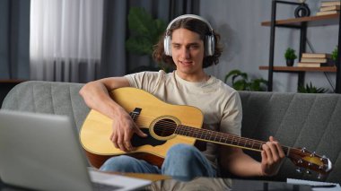 Günlük yaşam tarzı konsepti. Genç adam evde vakit geçiriyor, akustik gitarla kanepede oturuyor, kulaklık takıyor, dizüstü bilgisayarında şarkı söylüyor ve müzik dinliyor..