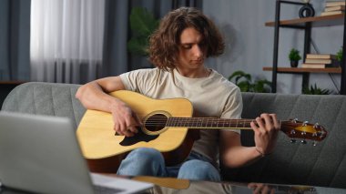 Günlük yaşam tarzı konsepti. Genç bir adam evde vakit geçiriyor, akustik gitarla kanepede oturuyor, bilgisayarla oynuyor ve kullanıyor, internetten öğreniyor..