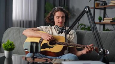 Günlük yaşam tarzı konsepti. Genç bir adam evde vakit geçiriyor, akustik gitarla kanepede oturuyor, akıllı telefondan canlı video kaydediyor, şarkı söylüyor ve çalıyor..