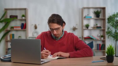 Günlük yaşam tarzı konsepti. Genç yetişkin bir adam evde vakit geçiriyor, erkek öğrenci masa başında oturuyor dizüstü bilgisayarla konuşuyor, video görüşmeleri yapıyor, not defteri yazıyor, ekranda notlar gösteriyor..