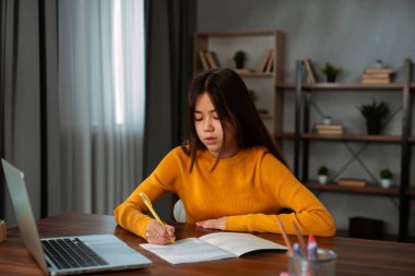 Sarı kazaklı genç bir kız masasında dizüstü bilgisayar kullanırken not defterine notlar yazmaya konsantre oluyor..