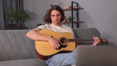 Günlük yaşam tarzı konsepti. Genç bir adam evde vakit geçiriyor, akustik gitarla kanepede oturuyor, gitar eşliğinde şarkı söylüyor, kafasını sallıyor, sürecin tadını çıkarıyor..