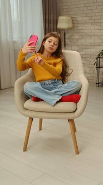 Günlük yaşam tarzı konsepti. Genç kız akşam vaktini evde geçiriyor, kız oturma odasında oturuyor ve akıllı telefondan selfie çekiyor, kameraya poz veriyor, ekrana bakıyor..