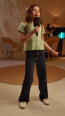 Günlük yaşam tarzı konsepti. Genç bir kız çocuğu akşamını evde geçirir, kız oturma odasında durur ve dans ederek zıplar, saç tarağıyla şarkı söyler, pozitif bir ifade takınır. Yavaş çekim, dikey video.