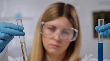 Güvenlik gözlüğü ve eldiven takan bir bilim adamı laboratuvar ortamında deney tüplerini inceliyor ve deneylere odaklanıyor. Kapat.