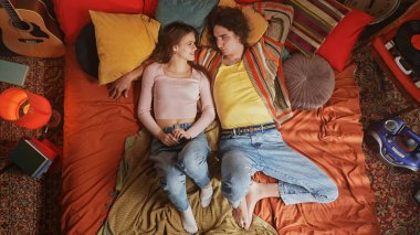 Renkli yastıklar ve retro dekorlarla çevrili turuncu bir yatakta birlikte huzurlu bir anın tadını çıkarıyorlar. Üst çekim